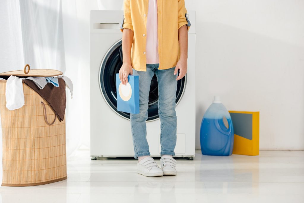 cropped view of child in yellow shirt and jeans holding washing powder in laundry room. Como cuidar tus prendas impresas con DTF. Viva DTF venta de transfer DTF por metros